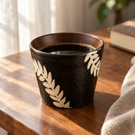 Zwarte keramische koffiekop met ivoren botanische takjes en zwarte koffie op een notenhouten tafel naast een boek en linnen doek.