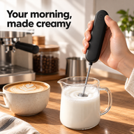 Black handheld milk frother used in a glass milk pitcher beside a latte at a home coffee station