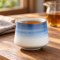 Blaue Verkostungstasse aus Keramik, mit Tee gefüllt, auf einem Holztisch neben einem Glas-Teekrug.