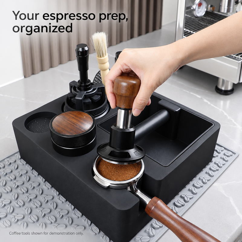 Hand tampt Kaffeemehl in einem Siebträger mit Holzgriff, gestützt von der schwarzen Espressostation; die Kaffeewerkzeuge dienen nur der Veranschaulichung und sind nicht enthalten.