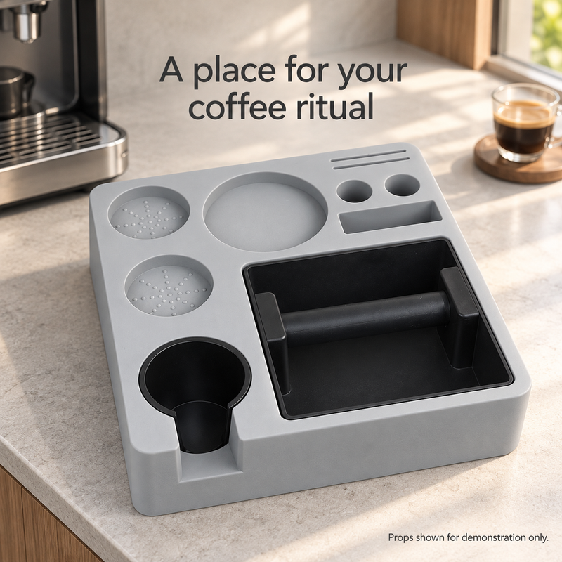Hellgrauer Espresso-Organizer mit schwarzer Abschlagbox und Halter neben einer Kaffeemaschine und Espressotasse auf einer Arbeitsfläche am Fenster.