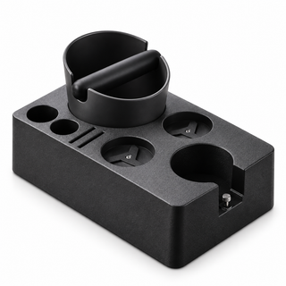 Front three-quarter view of a black espresso tamping station with empty tool holders, puck screen slots and a knock box on a pure white background.