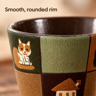 Close-up of the patchwork ceramic cup's rounded rim, brown glazed interior and painted cat and house motifs.