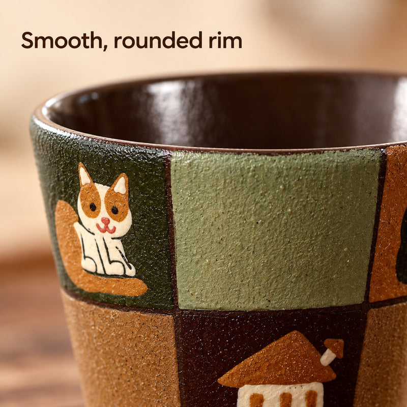 Close-up of the patchwork ceramic cup's rounded rim, brown glazed interior and painted cat and house motifs.