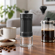 Ręczny młynek żarnowy Yozcoffee ze szklanym pojemnikiem obok french pressa i filiżanki kawy