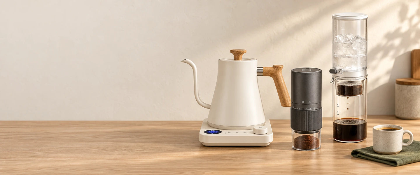 Gooseneck kettle, electric coffee grinder and glass cold drip coffee maker on a sunlit wooden coffee bar.